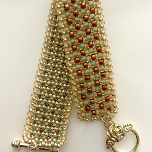 K&ouml;nnte beinhalten: Ein Armband aus Perlen in Gold-, Silber- und Pfirsichfarben. Das Armband hat einen rosenf&ouml;rmigen Verschluss und die Worte "Many Stitches" sind auf dem wei&szlig;en Hintergrund gedruckt.
