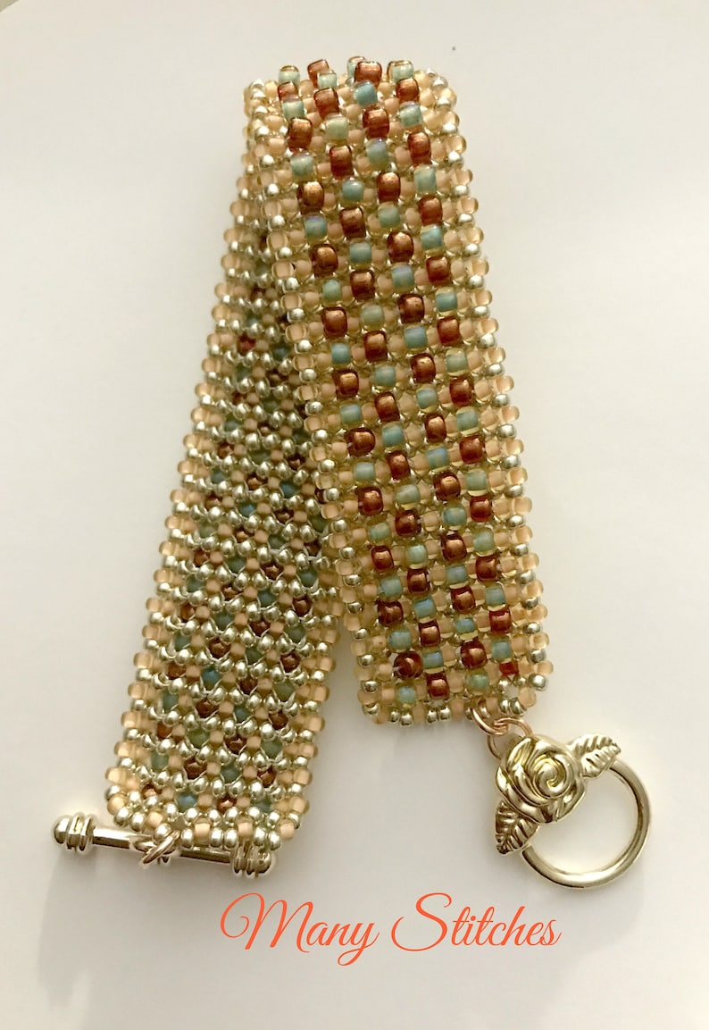 K&ouml;nnte beinhalten: Ein Armband aus Perlen in Gold-, Silber- und Pfirsichfarben. Das Armband hat einen rosenf&ouml;rmigen Verschluss und die Worte "Many Stitches" sind auf dem wei&szlig;en Hintergrund gedruckt.