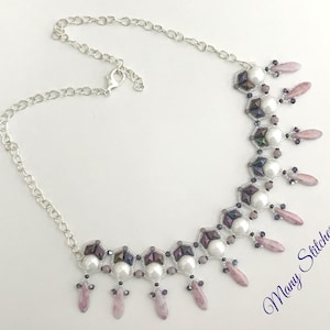Puede incluir: Un collar de cadena plateada con un colgante de cuentas que presenta perlas blancas, cuentas iridiscentes de color morado y rosa, y piedras de color rosa en forma de lágrima. El colgante tiene un diseño geométrico con un patrón repetitivo. El texto "Many Stitches" es visible en la esquina inferior derecha de la imagen.