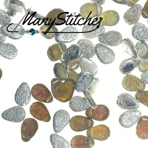 May include: A collection of small, teardrop-shaped glass beads in various colors, including silver, brown, and clear. The beads have a textured surface and are scattered on a white background.