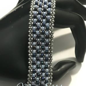 K&ouml;nnte beinhalten: Ein Armband aus Perlen in Silber und Blau. Das Armband besteht aus kleinen, runden Perlen in einem geometrischen Muster. Das Armband befindet sich auf einer schwarzen Mannequin-Hand.