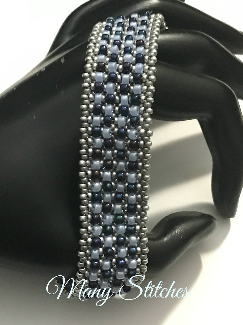 K&ouml;nnte beinhalten: Ein Armband aus Perlen in Silber und Blau. Das Armband besteht aus kleinen, runden Perlen in einem geometrischen Muster. Das Armband befindet sich auf einer schwarzen Mannequin-Hand.