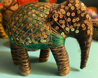 May include: A small, green and brown elephant figurine with gold sequins. The elephant is made of fabric and thread and has a detailed design.
