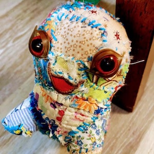 Può includere: Un giocattolo di stoffa colorata in patchwork con grandi occhi rotondi rossi e un sorriso cucito. Il giocattolo ha un cappello di stoffa verde ed è fatto di vari tessuti a motivi.