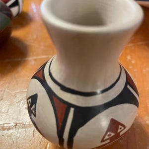 Puede incluir: Un pequeño jarrón de cerámica blanca con un patrón geométrico en negro, rojo y blanco. El jarrón tiene una boca ancha y un cuerpo redondeado.