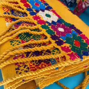 May include: A yellow woven fabric with colorful embroidered flowers and fringe. The fabric is folded and has a rope tied around it.