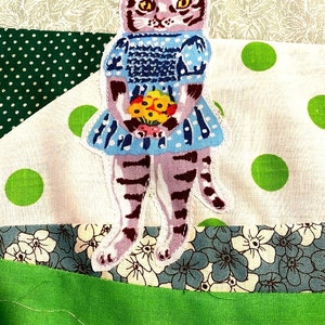 May include: A purple and brown striped cat wearing a blue dress with white polka dots and holding a bouquet of yellow flowers. The cat is standing on a patchwork fabric background.