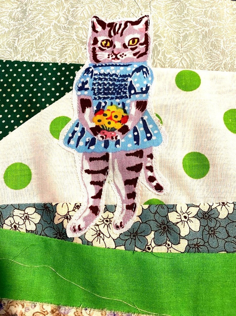 May include: A purple and brown striped cat wearing a blue dress with white polka dots and holding a bouquet of yellow flowers. The cat is standing on a patchwork fabric background.