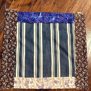 May include: A square patchwork quilt block with a blue and white striped fabric in the centre. The block is surrounded by a brown floral fabric, a purple floral fabric, and a beige floral fabric.