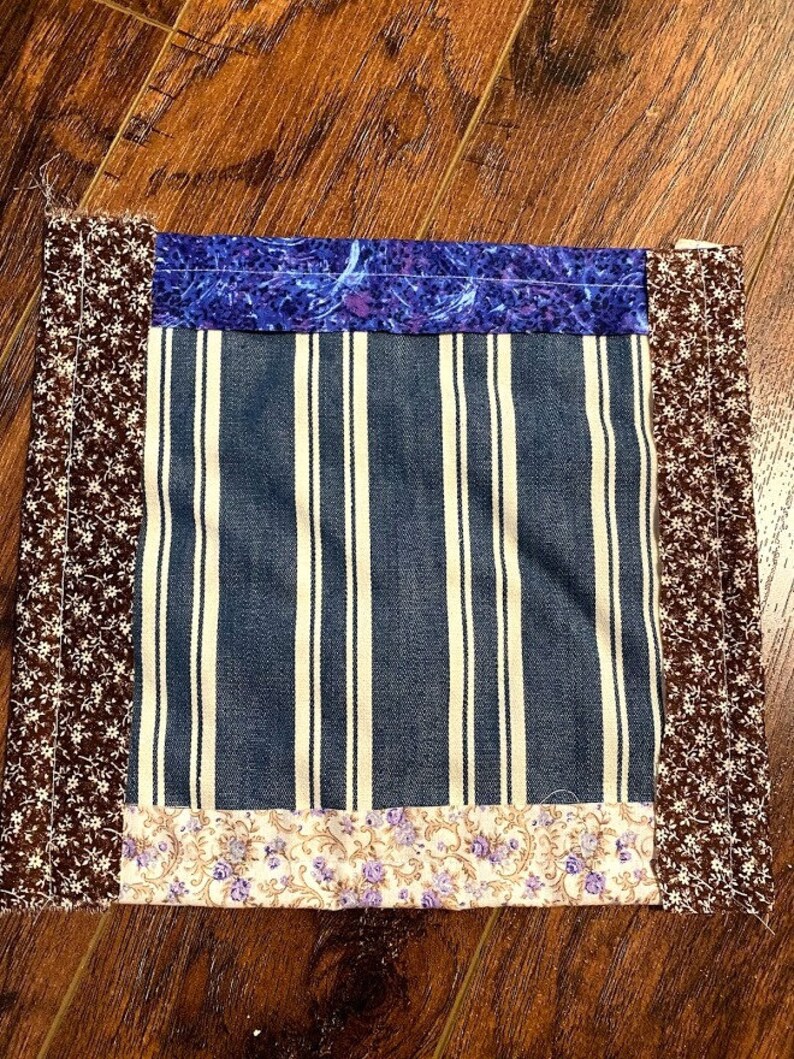 May include: A square patchwork quilt block with a blue and white striped fabric in the centre. The block is surrounded by a brown floral fabric, a purple floral fabric, and a beige floral fabric.