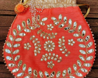 Puede incluir: Una bolsa de cordón de color coral con un diseño bordado en oro y plata. La bolsa tiene una forma redonda y un borla de cuentas.