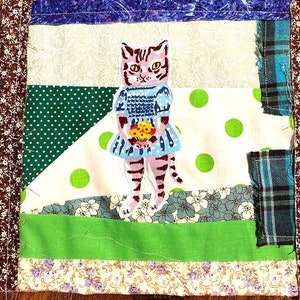 Könnte beinhalten: Ein Patchwork-Quilt-Quadrat mit einer handgenähten Katze, die ein blaues Kleid trägt und einen Blumenstrauß hält. Das Quadrat besteht aus verschiedenen Stoffen mit grünen, weißen und blauen Punkten, Blumenmustern und kariertem Stoff.
