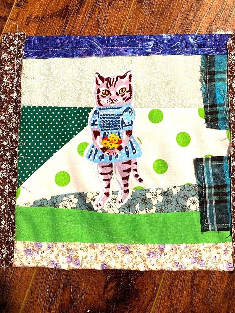 May include: A patchwork quilt square featuring a hand-stitched cat wearing a blue dress and holding a bouquet of flowers. The square is made of various fabrics with green, white, and blue polka dots, floral patterns, and plaid.