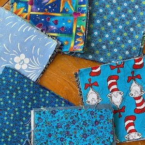May include: Assortment of blue fabric squares with various patterns, including a sun with a face, a floral design, a small blue and green polka dot pattern, a blue and white floral pattern, and a blue pattern with a cat wearing a red and white hat.