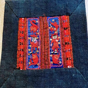 Pode incluir: Um quadrado de tecido azul escuro com um desenho bordado em vermelho, azul e dourado. O desenho apresenta duas figuras com roupas vermelhas e detalhes dourados, cercadas por peixes e outros padrões geométricos.