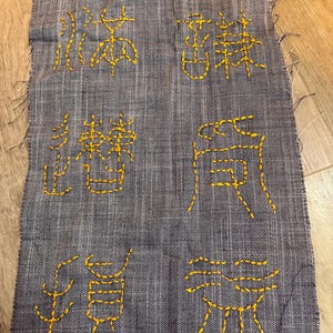 Puede incluir: Un trozo rectangular de tela gris con caracteres bordados en amarillo. La tela tiene una textura tejida y bordes deshilachados. Los caracteres bordados están dispuestos verticalmente.
