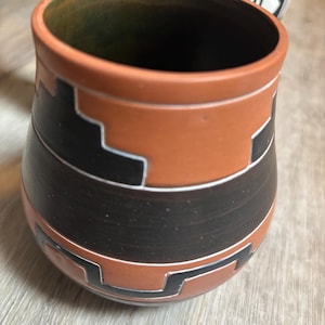 May include: A terracotta-colored ceramic cup with a rounded shape and a dark green interior. The exterior features a wide horizontal black band around the center and geometric step patterns in black with thin white outlines against the clay surface.