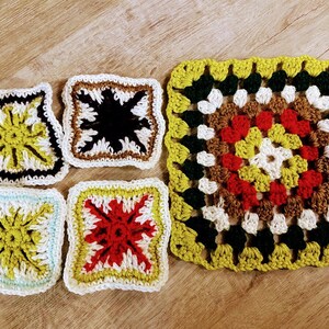 May include: Five crocheted squares in different colours and patterns. The squares are made with white, yellow, green, red, brown, and black yarn. The squares are arranged in a row on a wooden surface.