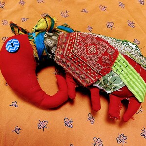 May include: A red fabric elephant toy with a blue button eye and patchwork fabric on its back. The elephant is lying on a patterned orange fabric.