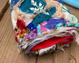 Könnte beinhalten: Eine zusammengerollte Patchwork-Decke aus verschiedenen Stoffen in den Farben Blau, Rot, Grün, Weiß und Gelb. Die Decke ist ausgefranst und hat einen Knopf an der Seite.