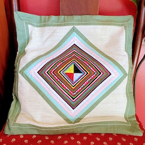 May include: A decorative pillow with a green border and a diamond-shaped design in the center. The diamond is made up of multiple layers of fabric in various colors, including pink, blue, green, yellow, red, and black.