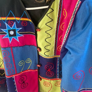 May include: A colorful patchwork jacket with a black background and a variety of geometric and floral embroidery. The jacket has a button closure and long sleeves. The label on the jacket reads "Julia Lee".