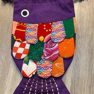 May include: Purple drawstring bag shaped like a fish with a patchwork design of colourful fabric. The fish has a large eye and a tail with white stitching.