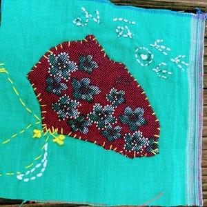 May include: A handmade textile art piece with a teal background. A maroon patch with floral embroidery is stitched onto the teal fabric. White and yellow thread accents the design, with text embroidered in white thread. The piece is on a wooden surface.