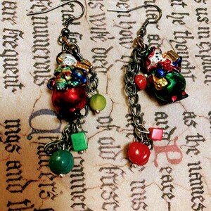 Può includere: Un paio di orecchini pendenti con un ciondolo a forma di pupazzo di neve con accenti rossi, verdi e dorati. Gli orecchini sono realizzati con una catena e varie perline colorate, tra cui rosso, verde e marrone.