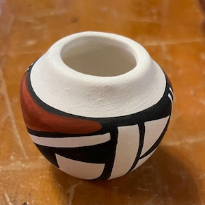 May include: A small, white ceramic pot with a geometric design in black, red, and white. The pot is round and has a wide opening.