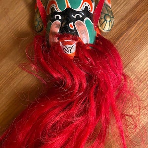 May include: A traditional Chinese opera mask with a green, black, and pink painted face, red horns, and a long, flowing red beard.