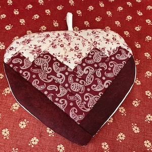 Puede incluir: Un agarrador de olla en forma de corazón con un patrón de paisley y floral. El agarrador de olla está hecho de tela acolchada y tiene un esquema de color burdeos, blanco y crema.
