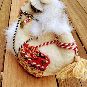 Könnte beinhalten: Weiche Plüschkamelfigur in Weiß mit einem rot-weiß gestreiften Sattel und goldenen Verzierungen. Das Kamel hat einen langen, flauschigen Schwanz und ein weißes Fell.