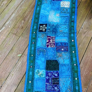 May include: A blue and green patchwork runner with embroidered details. The runner is made of various fabrics and has a fringed edge.