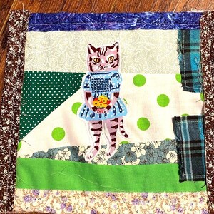 May include: A patchwork quilt square with a green, white and blue design. The centre of the square features an embroidered image of a cat wearing a blue dress and holding a bouquet of flowers.