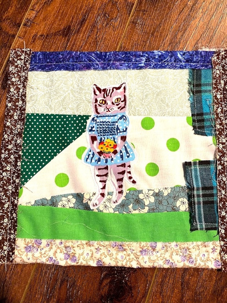 May include: A patchwork quilt square with a green, white and blue design. The centre of the square features an embroidered image of a cat wearing a blue dress and holding a bouquet of flowers.