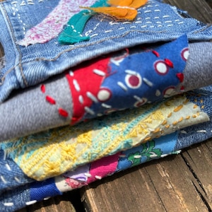May include: Close-up of a stack of denim fabric pieces with colorful fabric patches and decorative stitching. The patches include floral and abstract patterns in shades of pink, blue, yellow, and red. The denim is a light blue wash.