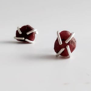 以下が含まれることがあります： 赤い宝石がそれぞれにセットされた、銀製のイヤリングのペア。宝石は不規則な形をしていて、ざらざらした質感です。銀は星形のデザインです。