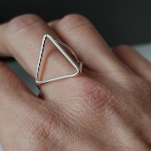 Könnte beinhalten: Ein silberner Dreiecksring mit einem schlichten, geometrischen Design. Der Ring wird an einem Finger getragen.