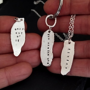 Könnte beinhalten: Drei silberne Anhänger mit gestempeltem Text. Einer lautet "snap out of it", ein anderer "you are the one" und der dritte "f l e r o e". Die Anhänger sind an einem Schlüsselanhänger und zwei Halsketten befestigt.