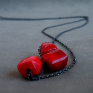 Può includere: Una collana a catena nera con tre perle di corallo rosso. Le perle sono di forma quadrata e hanno una finitura lucida.
