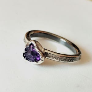 May include: A silver ring with a dark band and a heart-shaped, deep purple amethyst gemstone. The ring has a textured, antique finish, and the gemstone is set in a silver bezel.