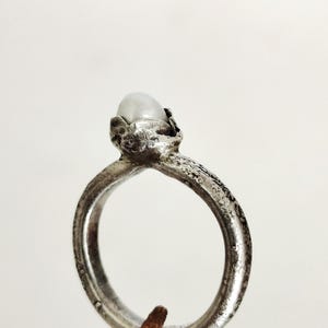 May include: A silver ring featuring a textured band and a small, oval, pale stone. The ring's setting has an intricate, organic design. The ring is shown against a plain white backdrop.