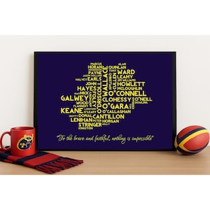 May include: A framed print with a navy blue background features a yellow text design in the shape of a map. The text includes names and phrases. A red mug and a striped scarf are on the left, and a red and yellow ball is on the right.
