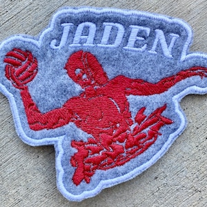 May include: A grey felt patch with white stitching. The patch features a red embroidered image of a person playing water polo, with the name "Jaden" embroidered in white above the image.