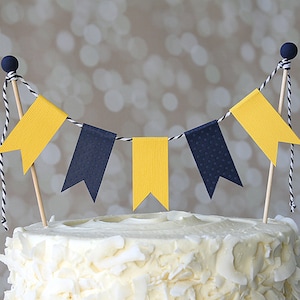 May include: A white cake with a yellow and navy blue paper banner on top. The banner is made of small flags with a string connecting them.