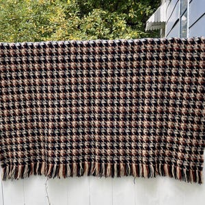 May include: A rectangular houndstooth patterned blanket in shades of brown, black, and beige, with a fringed edge. The blanket is hanging on a white fence, with green foliage in the background.