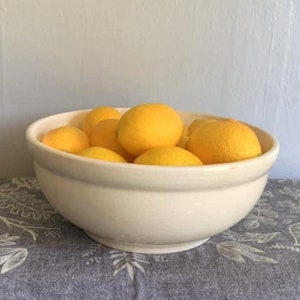 May include: A white ceramic bowl filled with yellow lemons. The bowl is sitting on a gray and white patterned tablecloth.