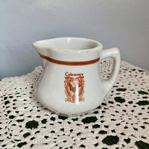 May include: A small, white ceramic creamer with a brown band around the rim and a handle. The creamer features the text "Coleman's" and a brown emblem. The creamer is on a white lace surface.