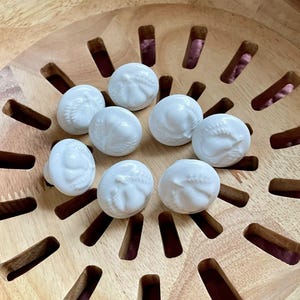May include: A collection of eight white ceramic decorative spheres, each with a detailed embossed design, arranged in a wooden bowl with a slotted pattern. The spheres are evenly spaced, creating a visually appealing arrangement.